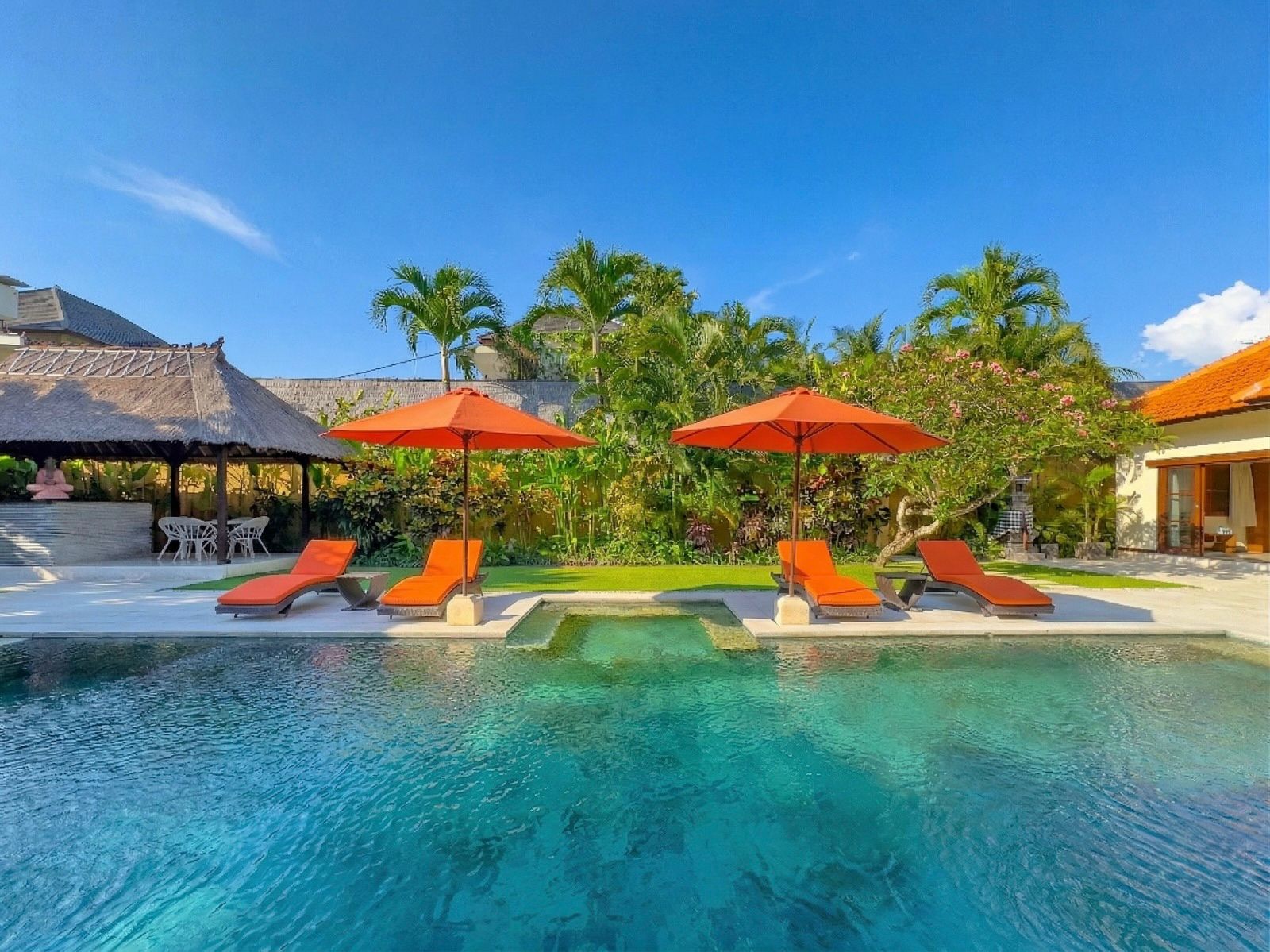 Bright daytime view of the main poolside deck at Villa Vara Seminyak by Villa Bugis Seminyak villas, showcasing a clear blue pool surrounded by exotic greenery.